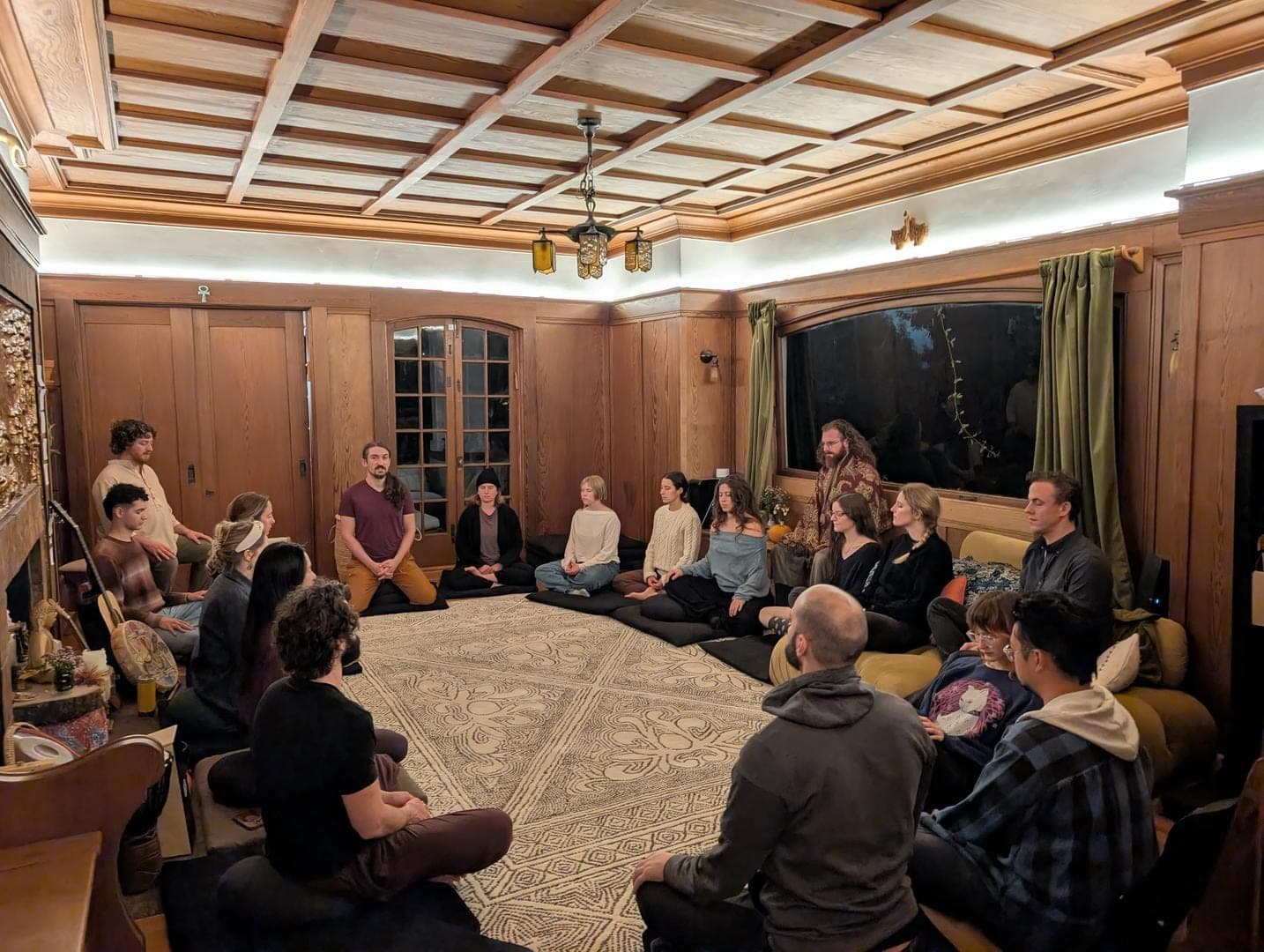 A group seated in a circle on cushions in a warm wood-paneled room, evening light
